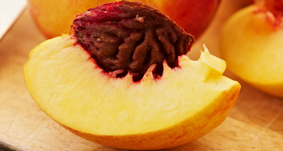 A sliced peach with a dark, wrinkled stone is presented on a wooden surface, with a blurred background of additional peaches.