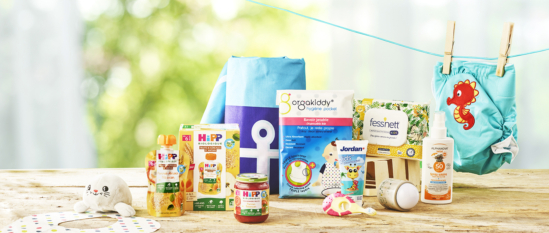 Divers produits et accessoires pour bébés, dont de la nourriture, des articles d'hygiène et un jouet en peluche, disposés sur une surface en bois avec un arrière-plan naturel flou.