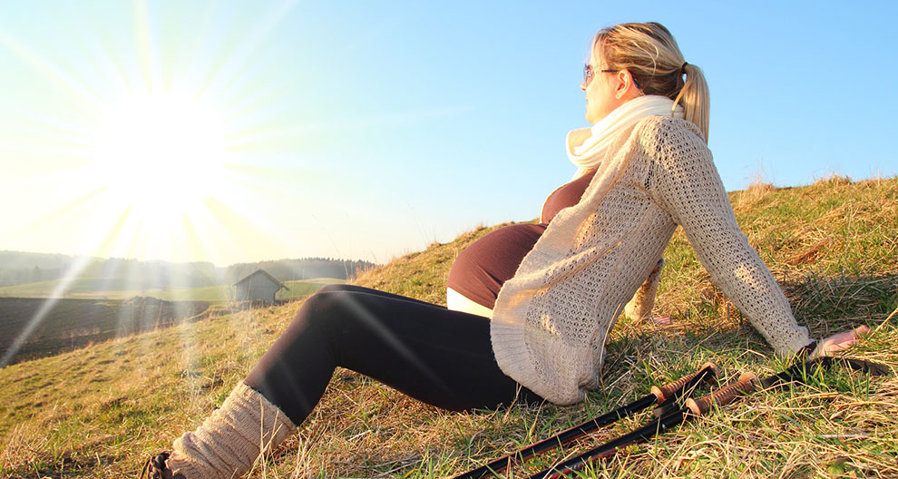 Une femme enceinte est assise sur une colline herbeuse, regardant le soleil, avec des bâtons de marche à côté d'elle.
