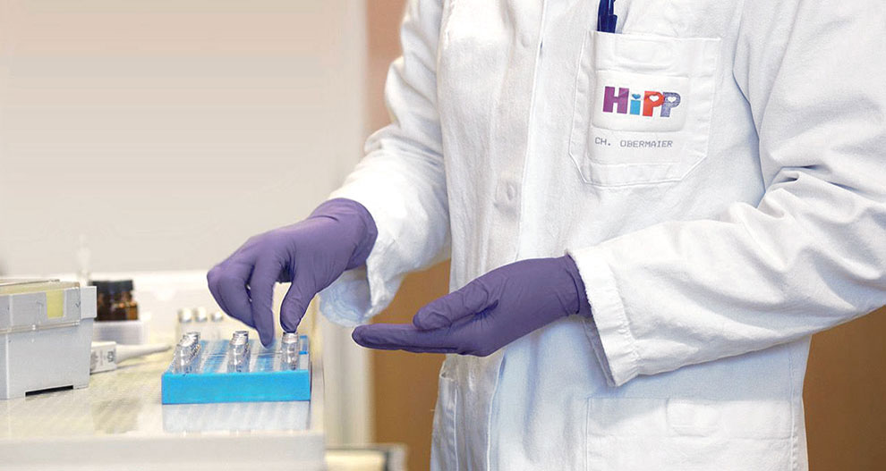 Une personne portant une blouse blanche avec le logo "HiPP" et le nom "CH. OBERMAIER" porte des gants violets et manipule un plateau bleu contenant plusieurs petits récipients.