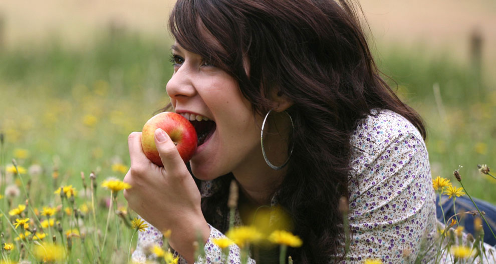 Une femme aux longs cheveux noirs est allongée sur l'herbe, entourée de fleurs jaunes, et croque une pomme rouge.