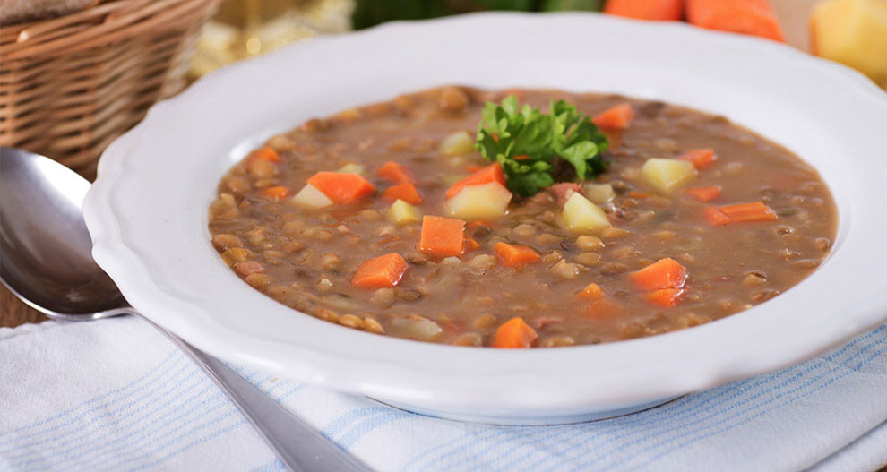Un bol de soupe épaisse et consistante avec des morceaux visibles de carottes, de pommes de terre et de lentilles est présenté sur une table avec une cuillère et une corbeille de pain à l'arrière-plan.