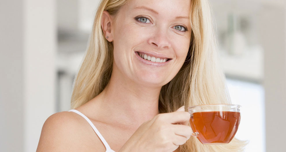 Une femme aux cheveux blonds tient une tasse en verre transparent remplie d'un liquide brun, peut-être du thé ou du café.