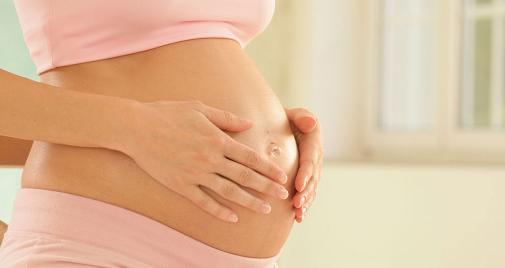 Une femme enceinte vêtue de rose, les mains délicatement posées sur son ventre, est assise à l'intérieur.