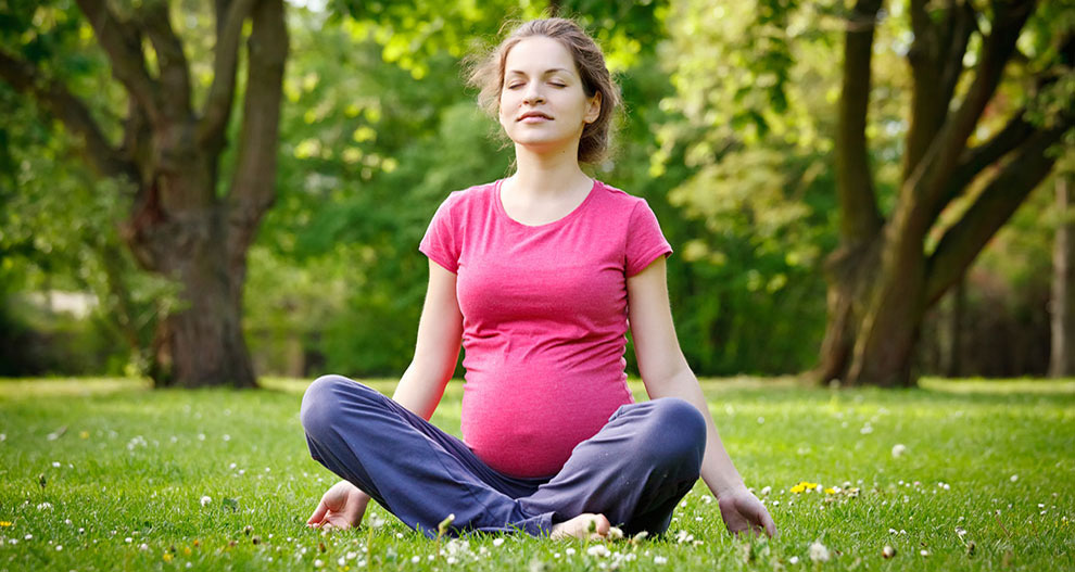 Une femme vêtue d'une chemise rose et d'un pantalon bleu est assise, les jambes croisées, sur un terrain herbeux et fleuri, entouré d'arbres.