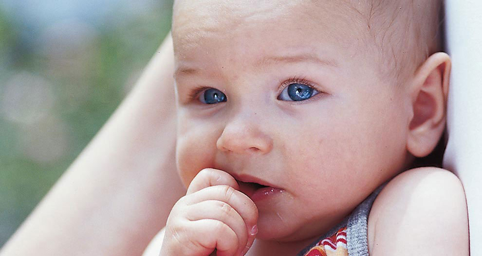 Un bébé aux yeux bleus repose sa tête sur l'épaule d'une personne, dont il mordille la main.
