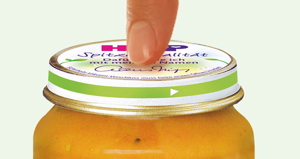 Une main s'apprête à ouvrir un pot de produit alimentaire de couleur orange avec un couvercle vert.