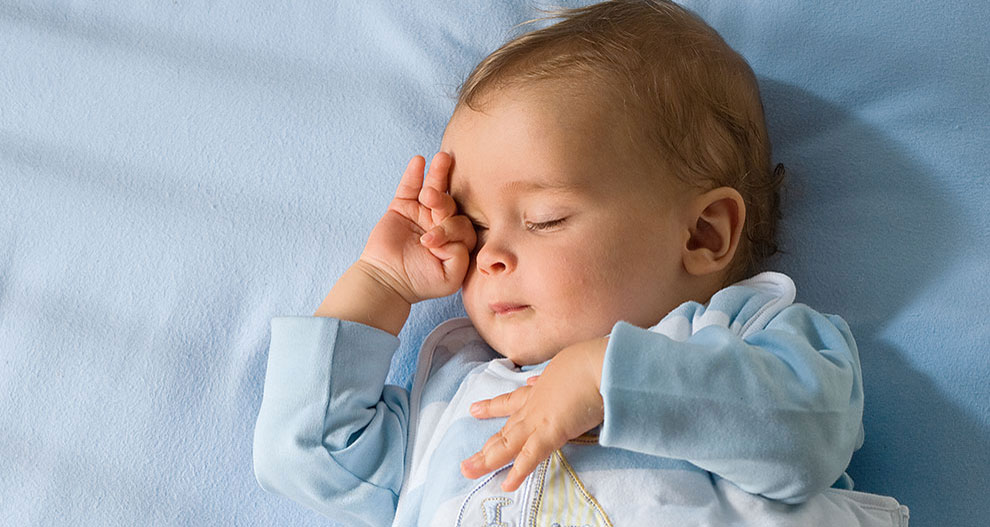 Un bébé est allongé sur un drap bleu. Il porte un vêtement bleu clair avec un motif de voilier et se couvre un œil avec une main.