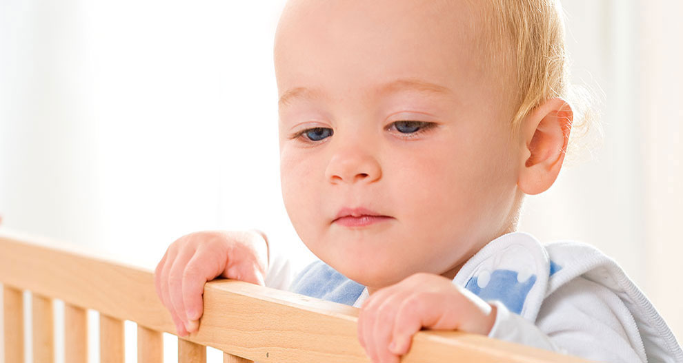 Un bébé aux cheveux clairs et aux yeux bleus regarde par-dessus la balustrade d'un berceau en bois.