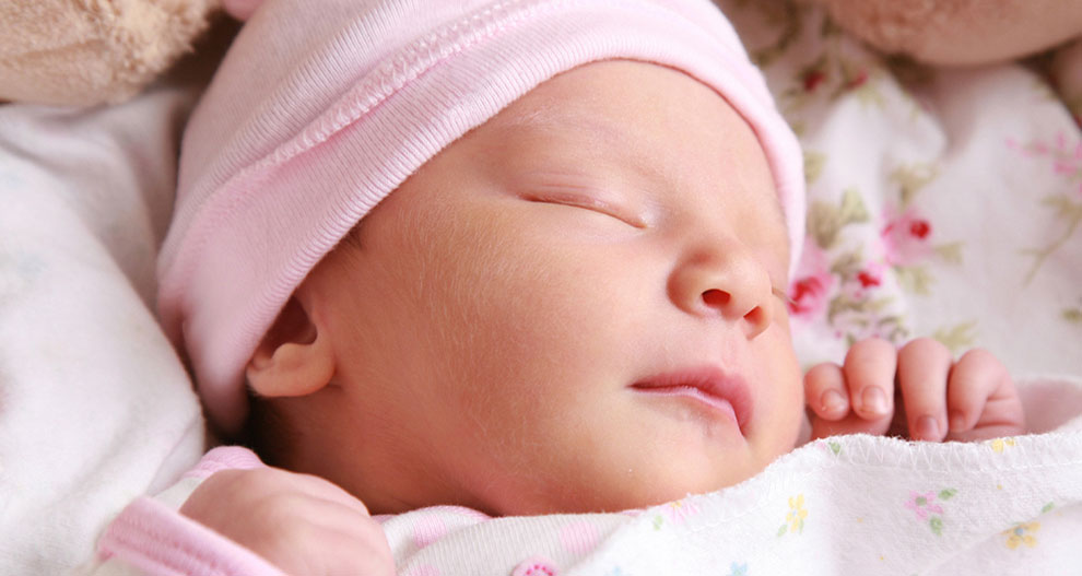 Gros plan sur le visage d'un bébé endormi, coiffé d'un bonnet rose et emmailloté dans une couverture blanche à motifs floraux.