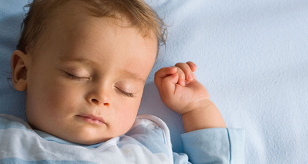 Un bébé est allongé sur un drap bleu, vêtu d'une chemise à rayures bleu clair et blanches, une main levée et les doigts recroquevillés.