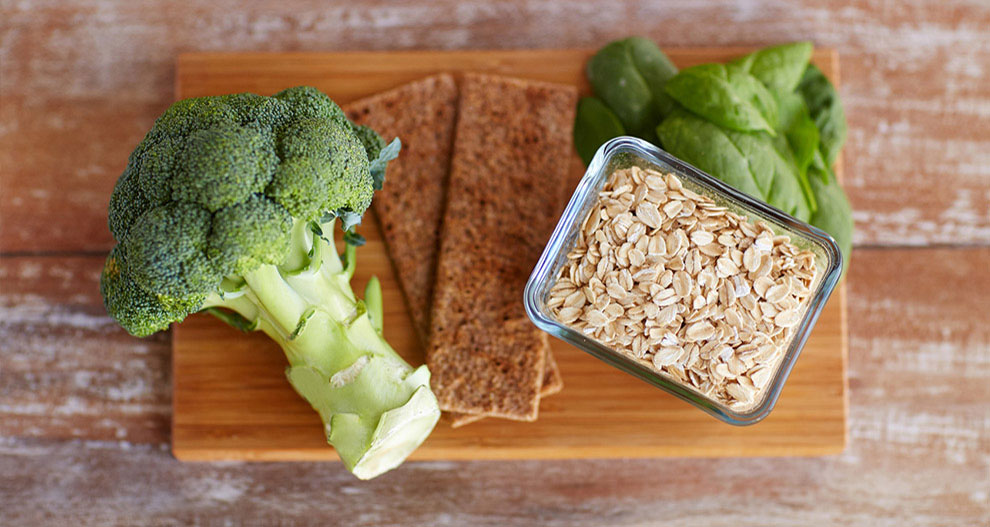 Une planche à découper en bois contient une tige de brocoli, un bouquet d'épinards frais, un récipient rectangulaire en verre rempli de flocons d'avoine et deux crackers bruns rectangulaires.