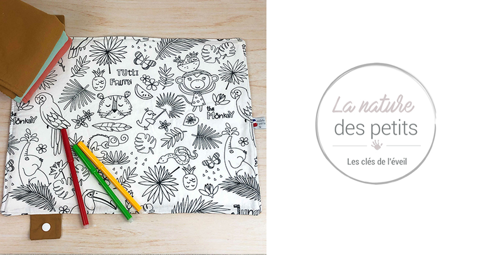 Un tapis en tissu avec des illustrations sur le thème de la jungle est accompagné de crayons de couleur et d'un classeur brun sur une surface en bois, avec les textes "La nature des petits" et "Les clés de l'éveil" dans un logo circulaire.