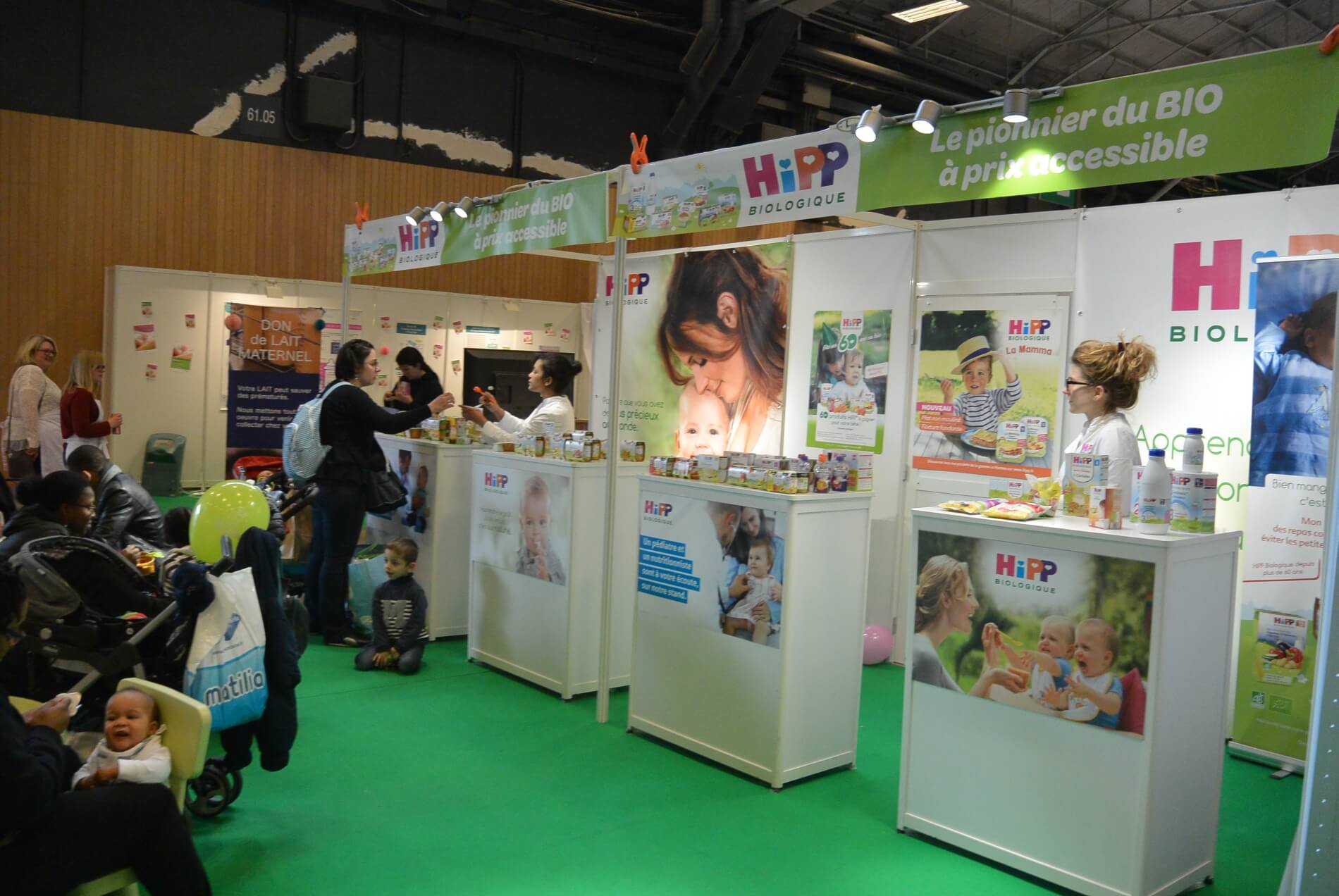Le stand d'HiPP au Salon Baby Un stand promouvant les produits pour bébés "HiPP Biologique" est au centre de l'attention, les participants interagissant avec l'exposition et les produits.